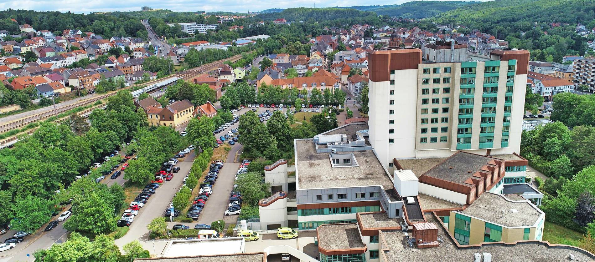 Panorama-Luftaufnahme einer Kleinstadt: rechts ein großes Klinikgebäude mit Hochhaus und grün getönten Fenstern, davor ein voller Parkplatz; links Bahngleise mit Bahnhof, dahinter Wohnhäuser und bewaldete Hügel unter leicht bewölktem Himmel.